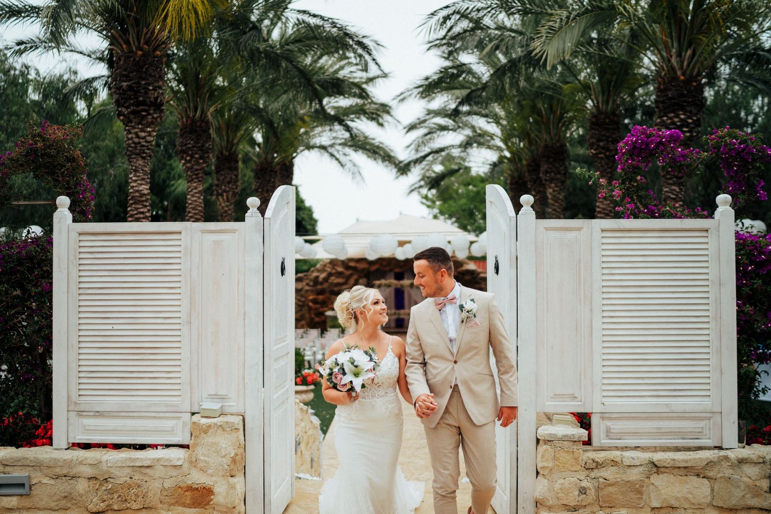 A romantic wedding couple stands between white gates surrounded by tropical palm trees and vibrant flowers.