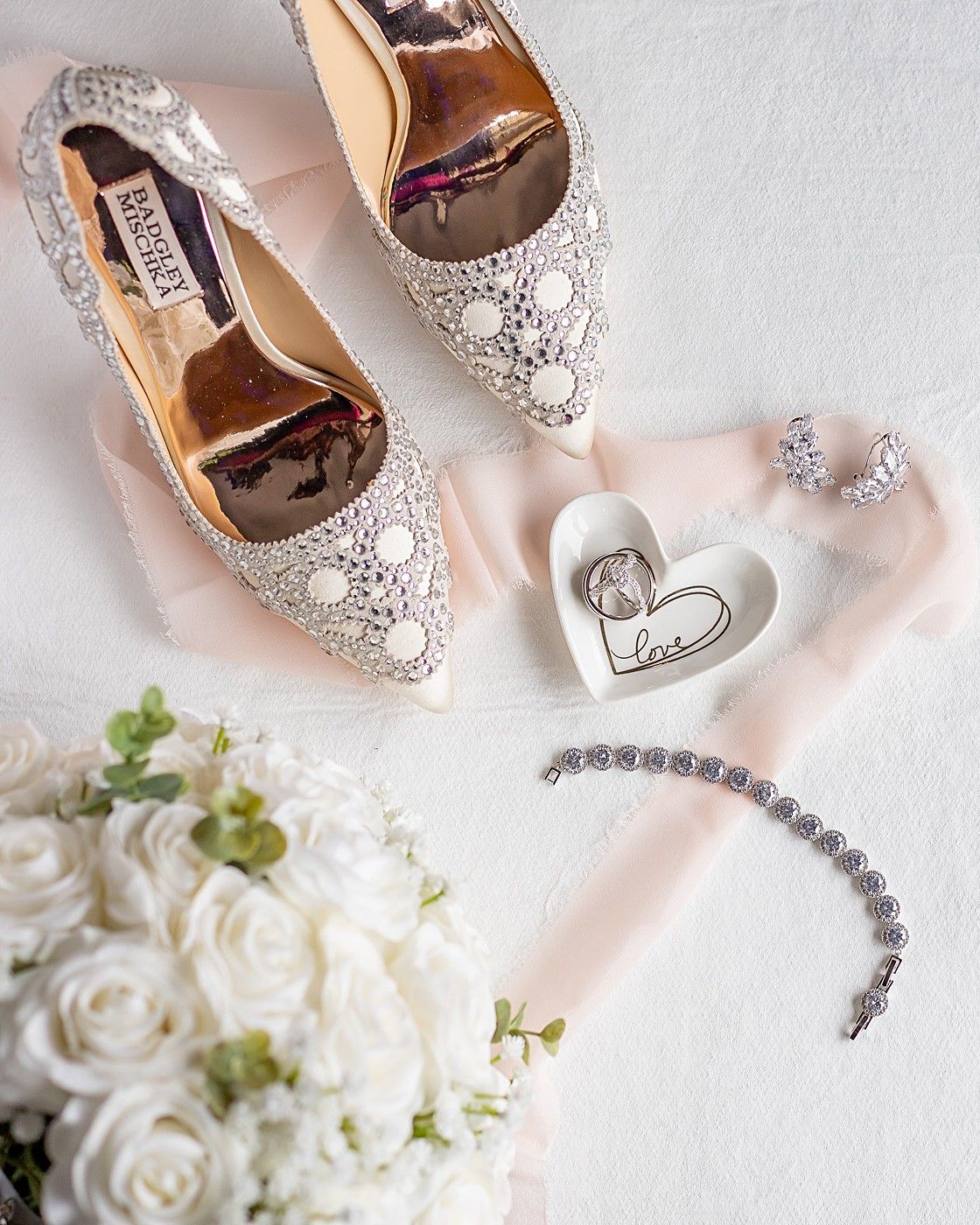 Bridal accessories flat lay with lace heels, white rose bouquet, diamond bracelet, and ring dish.