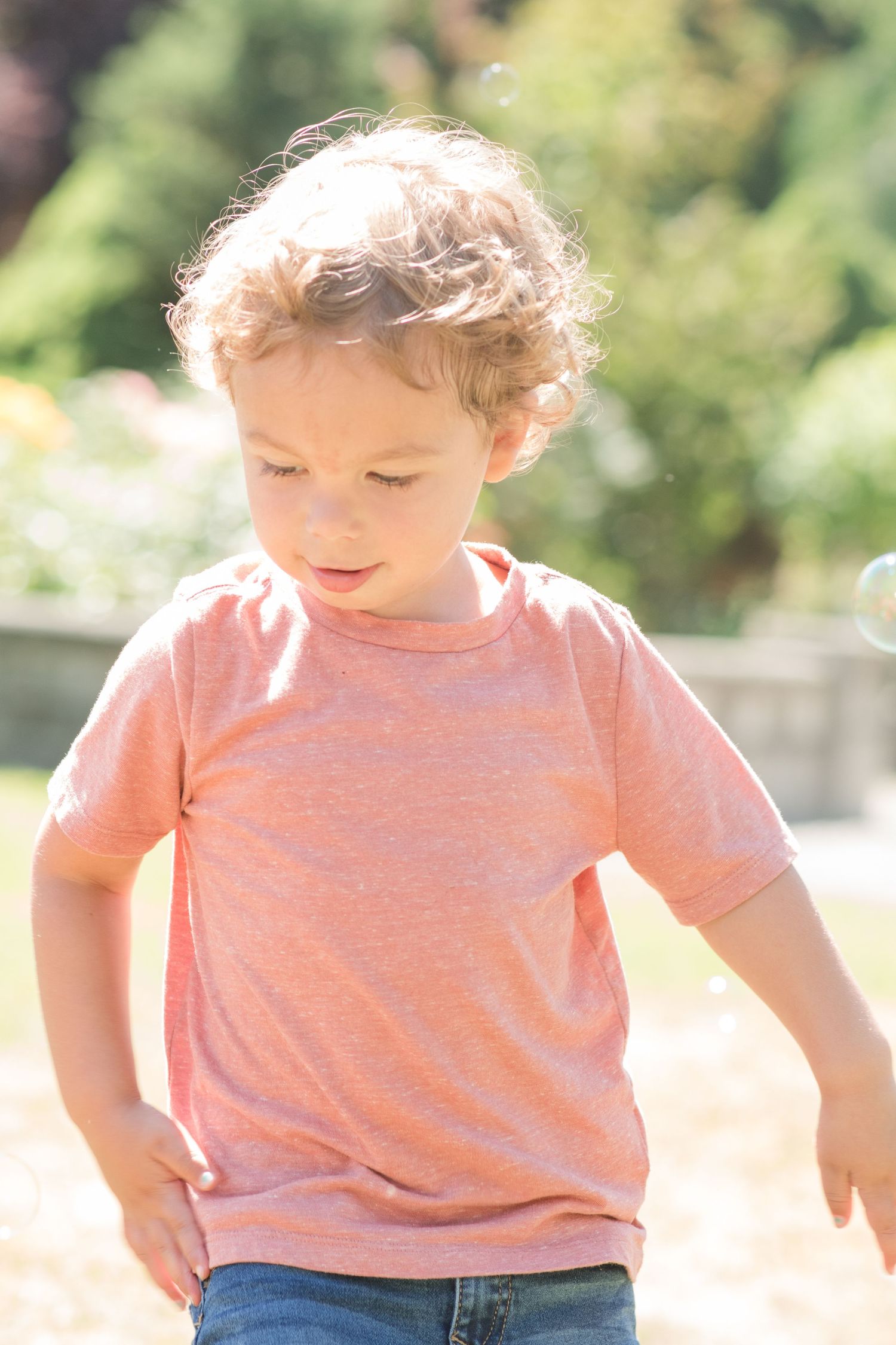 little boy with hair shining in the sun as he plays in the bubbles wearing a mauve shirt and looking down and away.