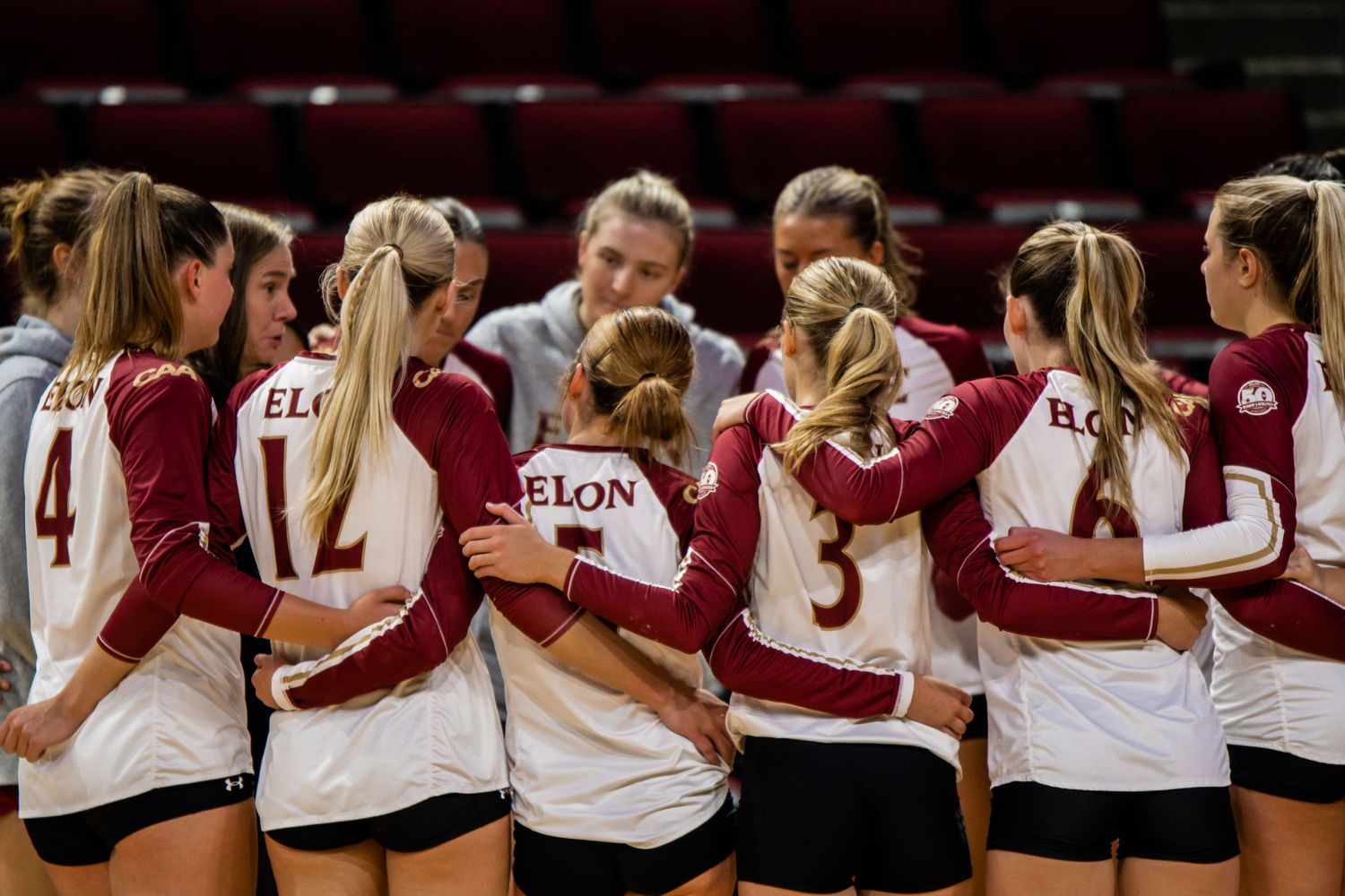 A volleyball team huddles together wearing maroon and white uniforms.