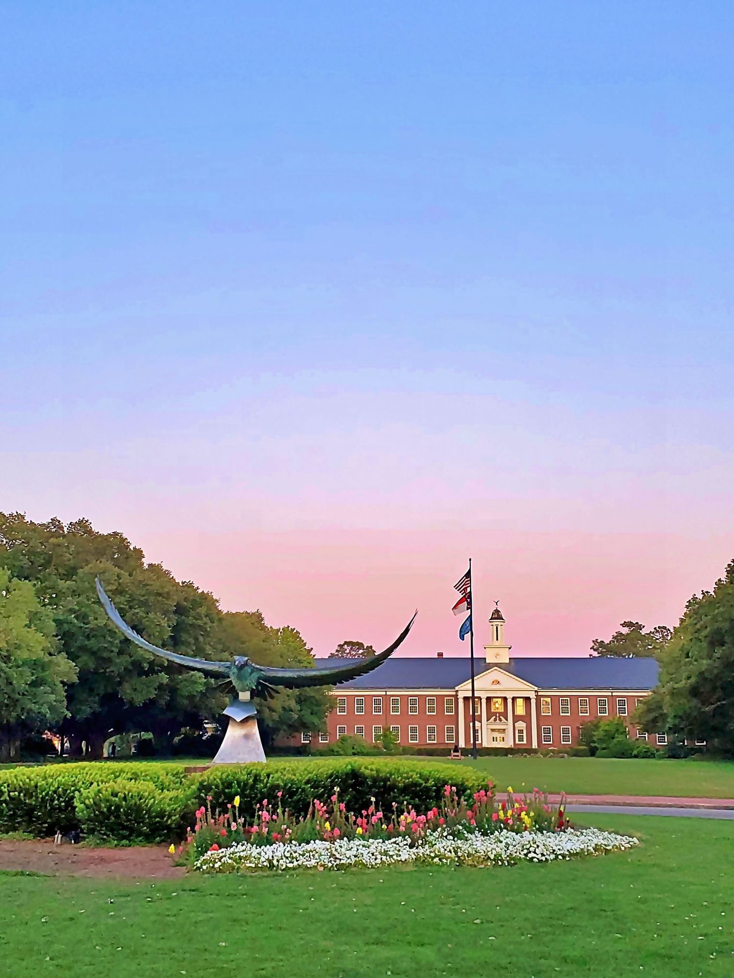 A beautiful campus building with a flower garden and statue at sunset.