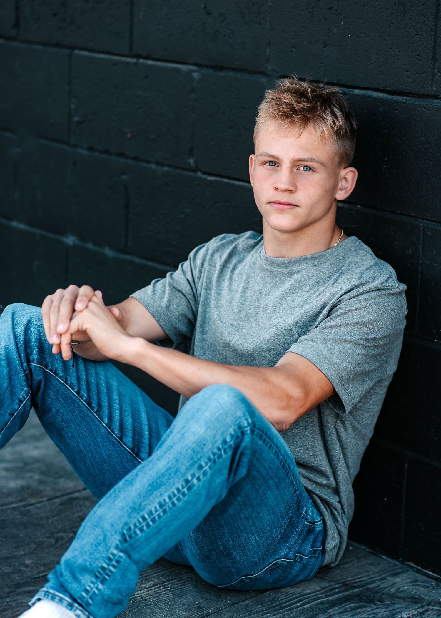 Casual portrait of student in grey shirt and jeans sitting against dark wall.