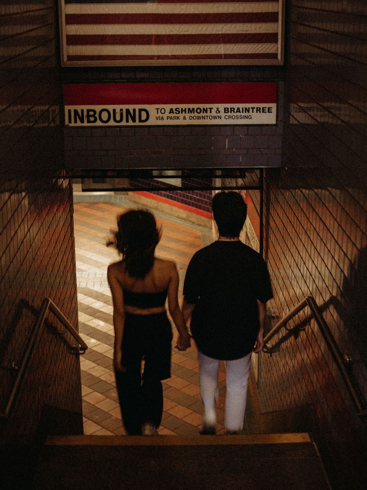 Two figures descending subway stairs under an American flag and Inbound sign.