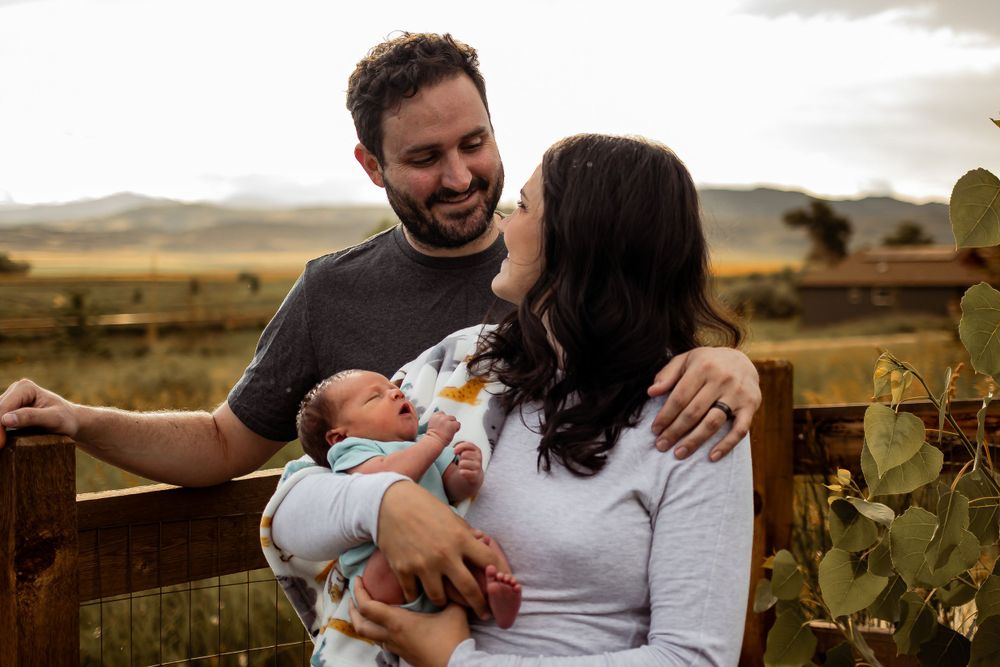 Family enjoys tender moment holding newborn baby against rustic wooden fence in golden countryside setting. Denver Family Photography.