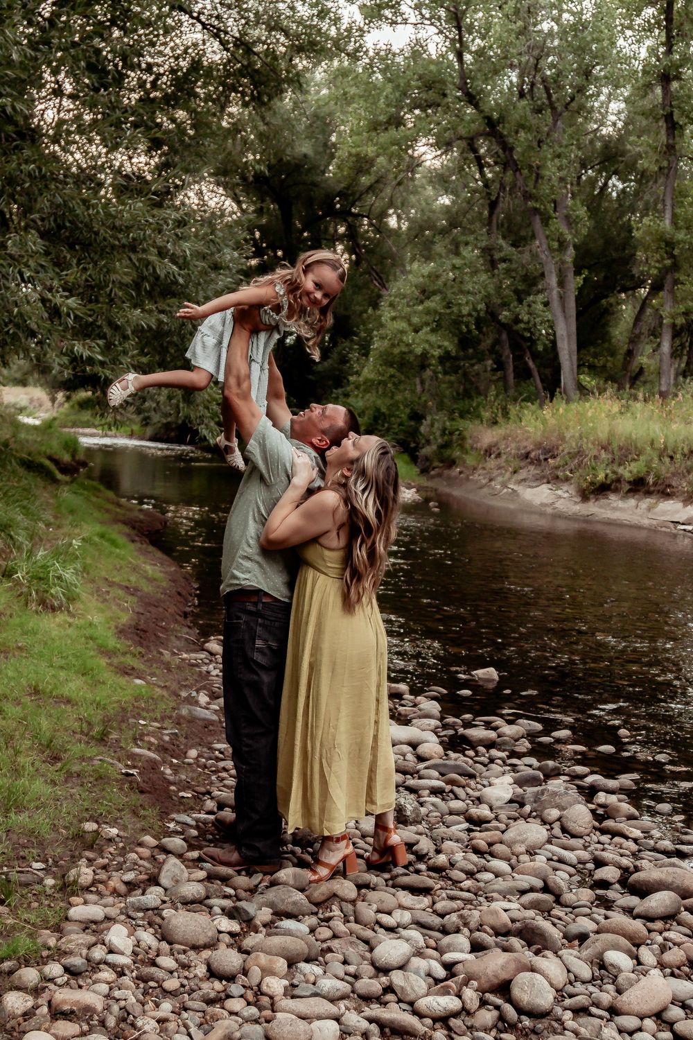 A couple plays with their child by a rocky stream in a wooded area. Denver Family Photography.