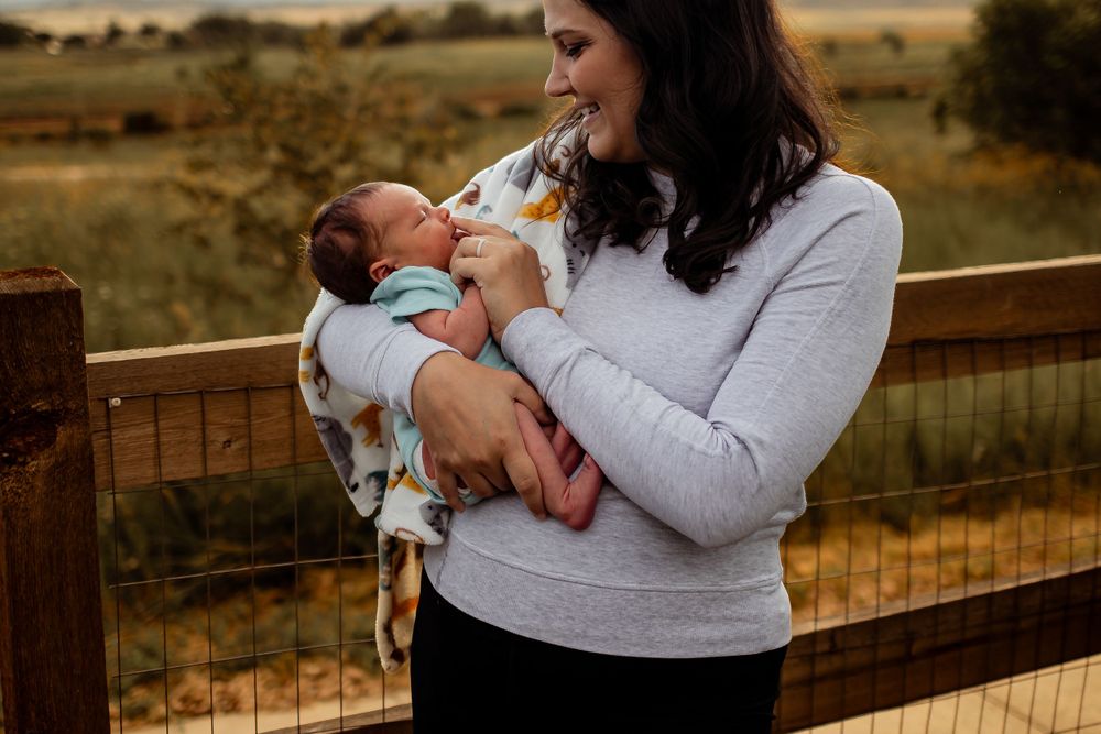 Mother in gray sweater cradles newborn baby while standing by wooden fence in rural landscape. Denver Family Photography.