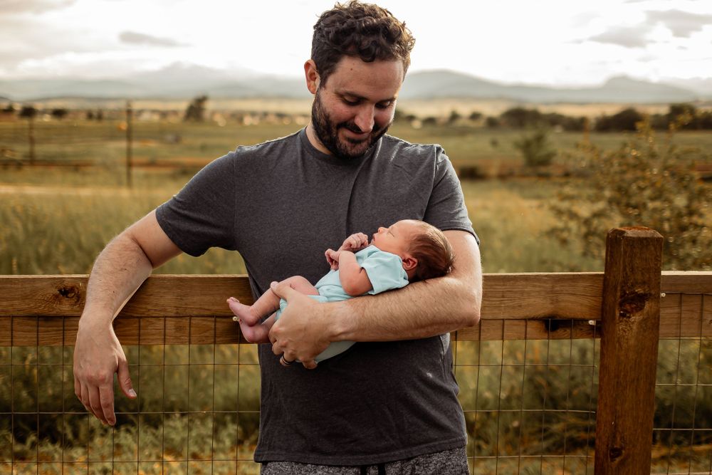 Parent holding sleeping newborn baby outdoors against rustic fence with scenic mountain backdrop. Denver Family Photography.