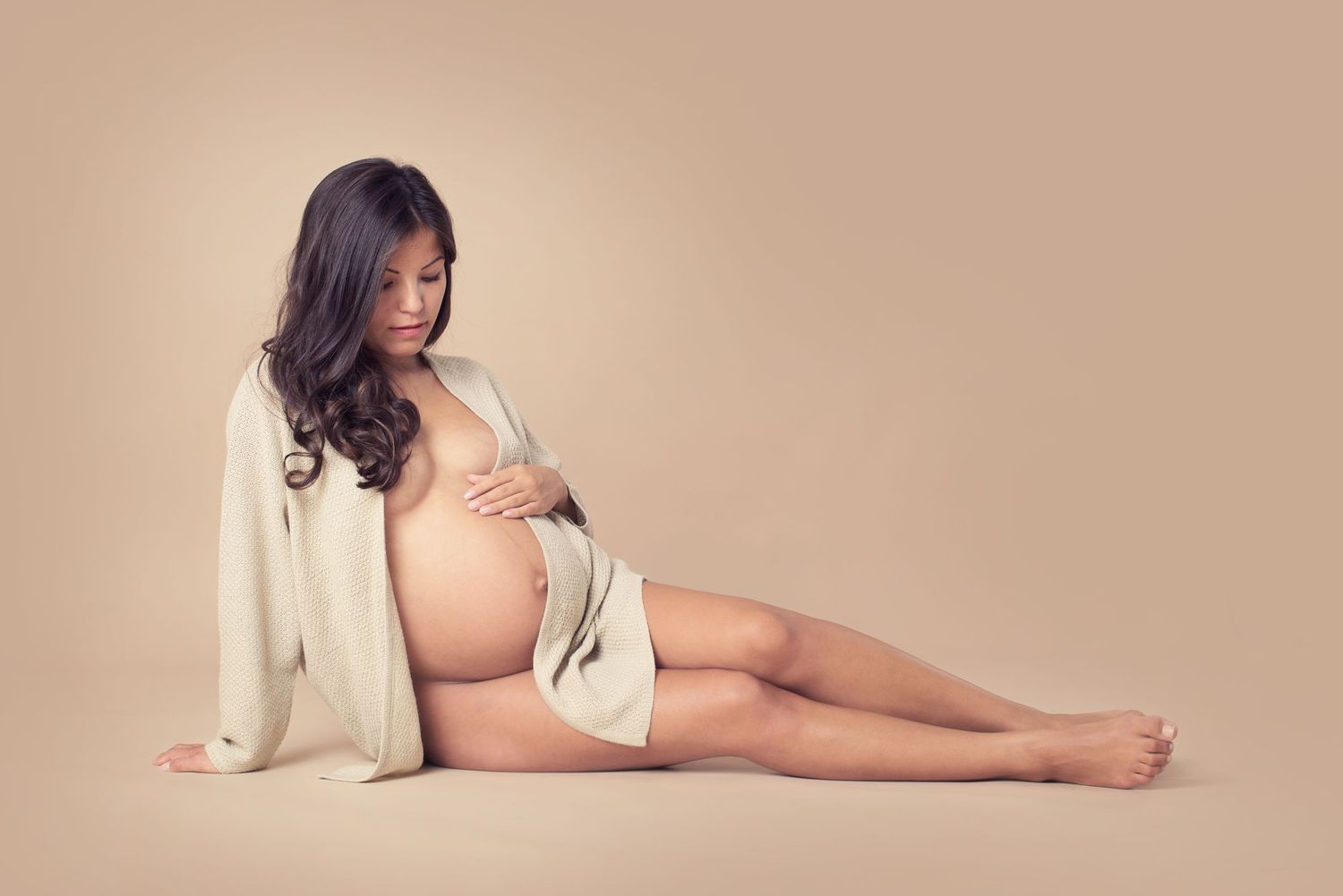 Maternity portrait of woman in white cardigan reclining against beige backdrop.