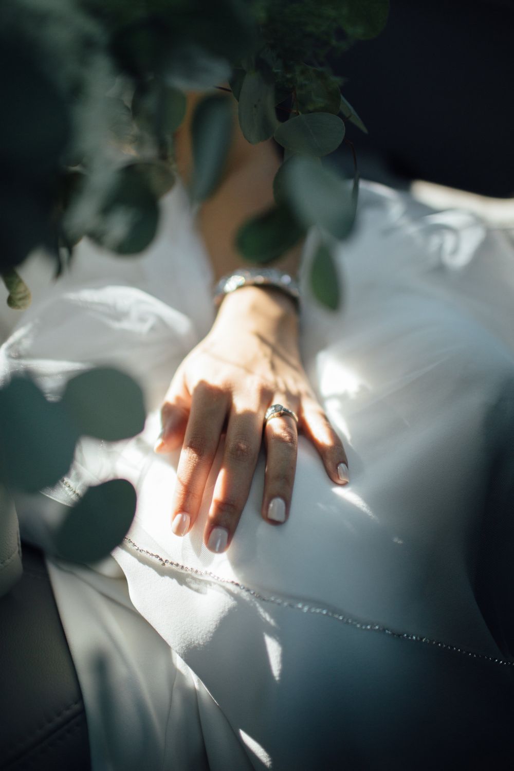 the hand of a bride sits on her knee, eucalyptus leaves in the foreground, her diamond engagement ring sparkling