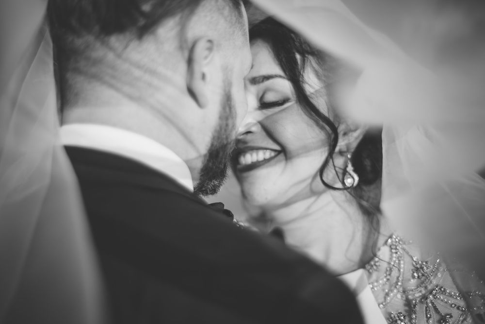 a bride smiling, eyes closed about to kiss her partner, photographer and couple are under her veil