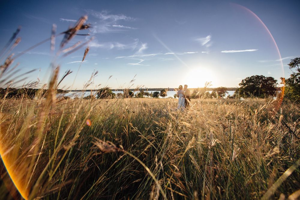 landscape photo, bright round sun flares in the corners, and a small bride & groom standing together in long grass