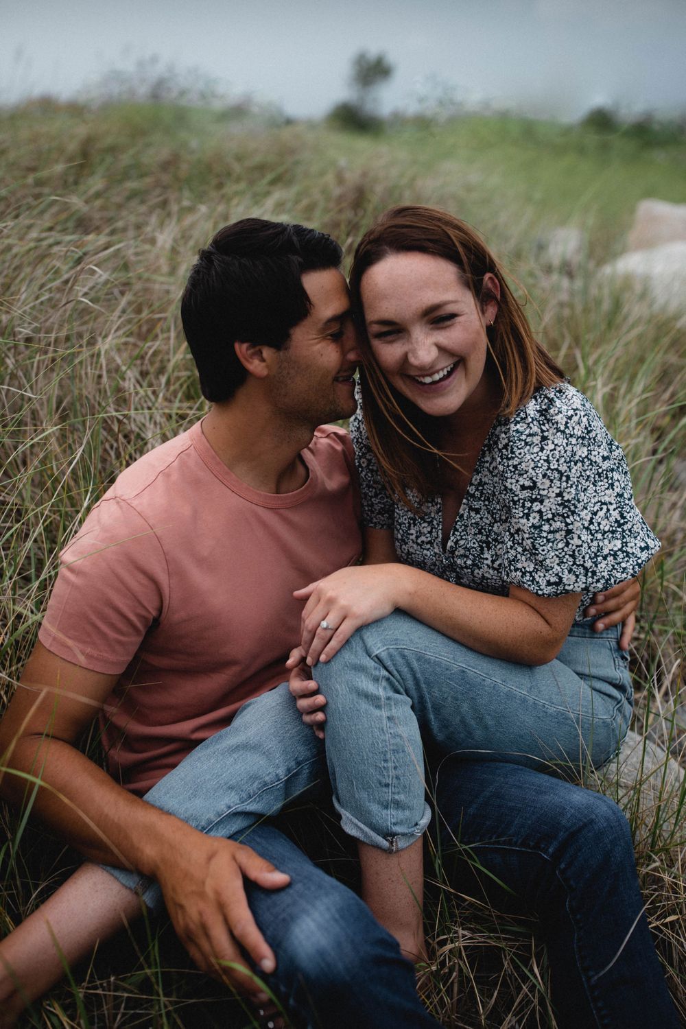 a white hetero couple sitting in the beach grass together. he whispers in her ear and she is laughing