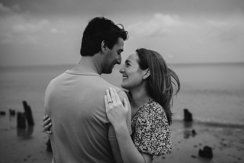 a black and white image of a white hetero couple, showing off her engagement ring on the beach