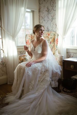 a bride in an old fashioned window lit chair holding a glass of champagne, with her dress spread out at her feet