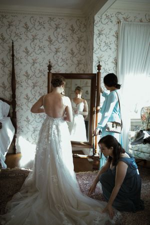 bride stands in an old fashioned room, looking in a large standing mirror, her friends adjust her dress and gaze at her