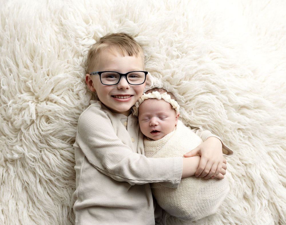bird's-eye view of smiling toddler boy with blonde hair and blue glasses with arms around newborn sister with headband