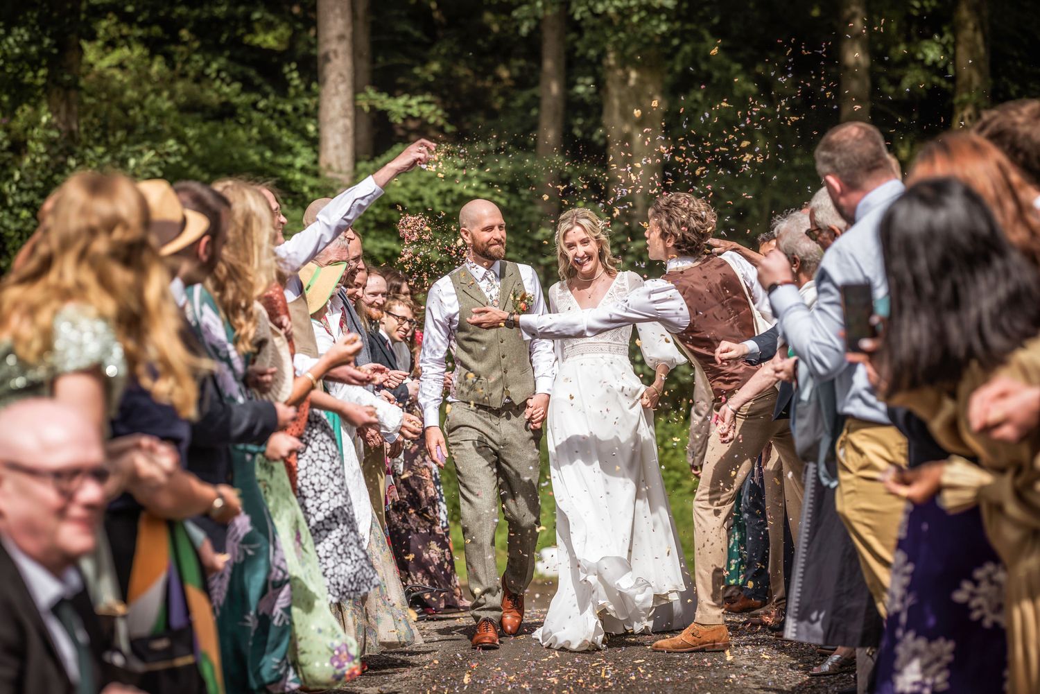 Joyful celebration with guests throwing confetti at couple walking through wooded path.