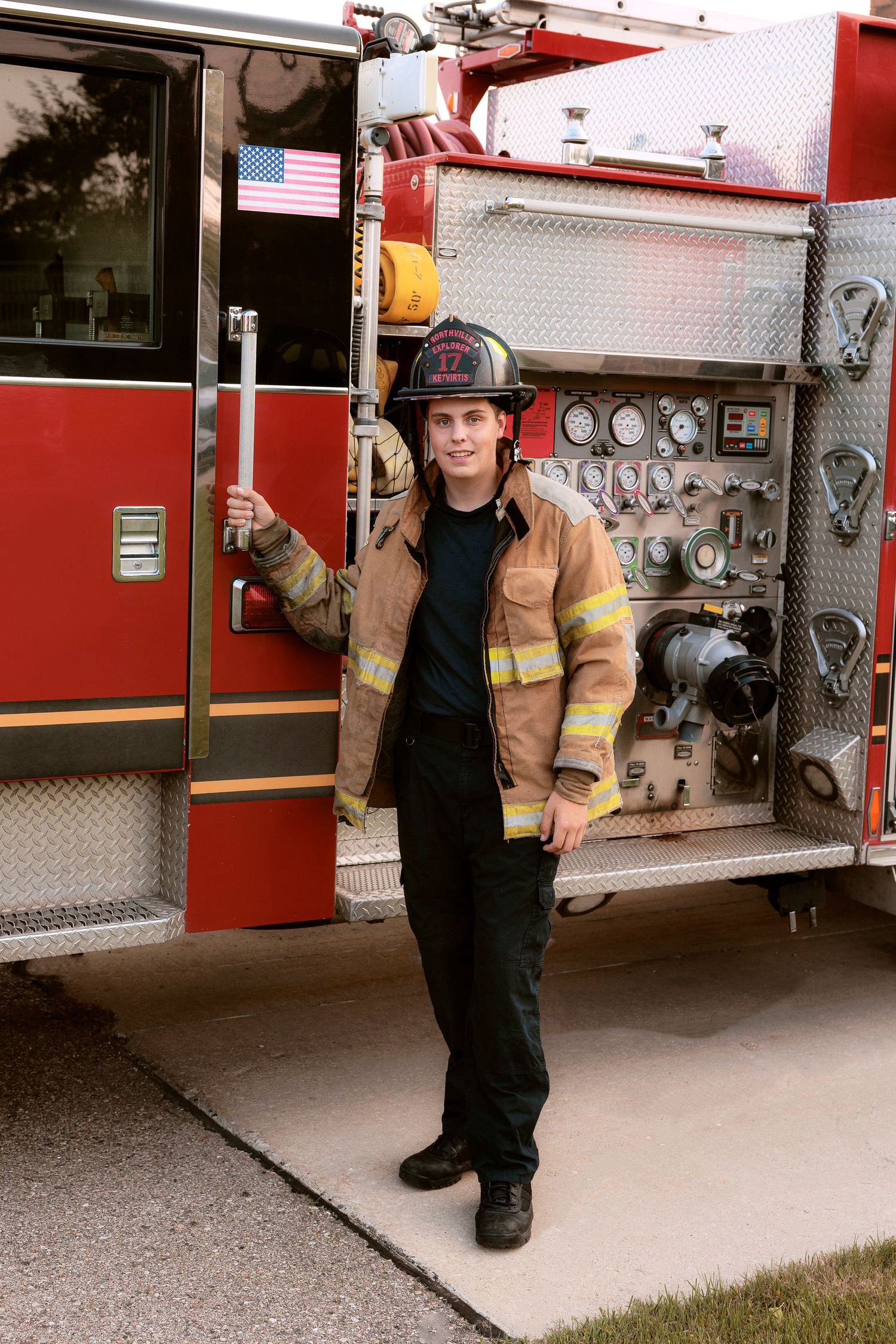 A firefighter in full gear poses beside a red fire engine wearing tan turnout gear.