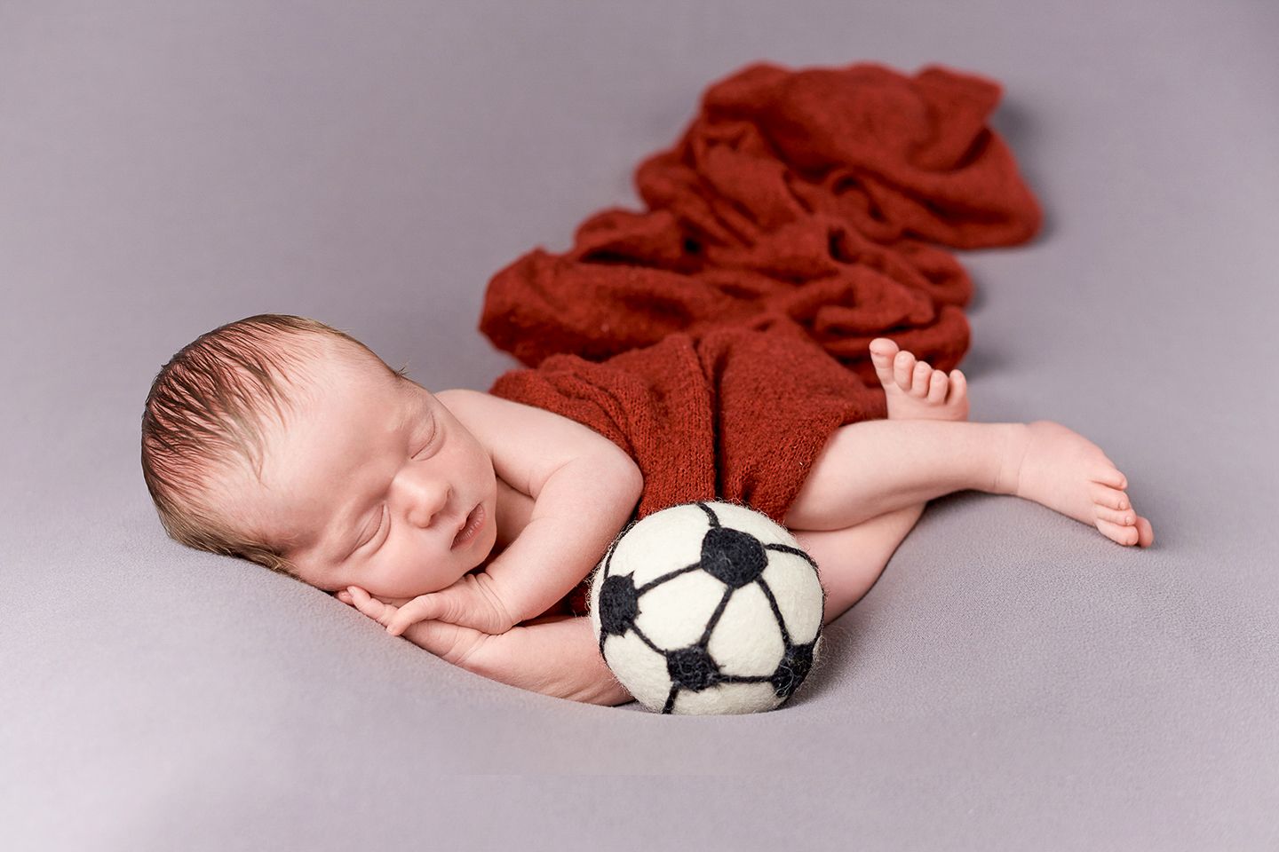 A sleeping newborn posed with a soccer ball and red fabric in a studio setting.