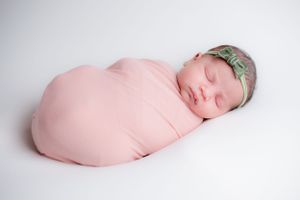 Sleeping newborn swaddled in soft pink fabric with mint green headband against white background.