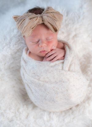 Peaceful newborn wrapped in cream lace swaddle with matching bow headband on fluffy white texture.