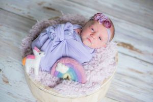 Newborn in purple outfit with rainbow plush toy resting in round basket.