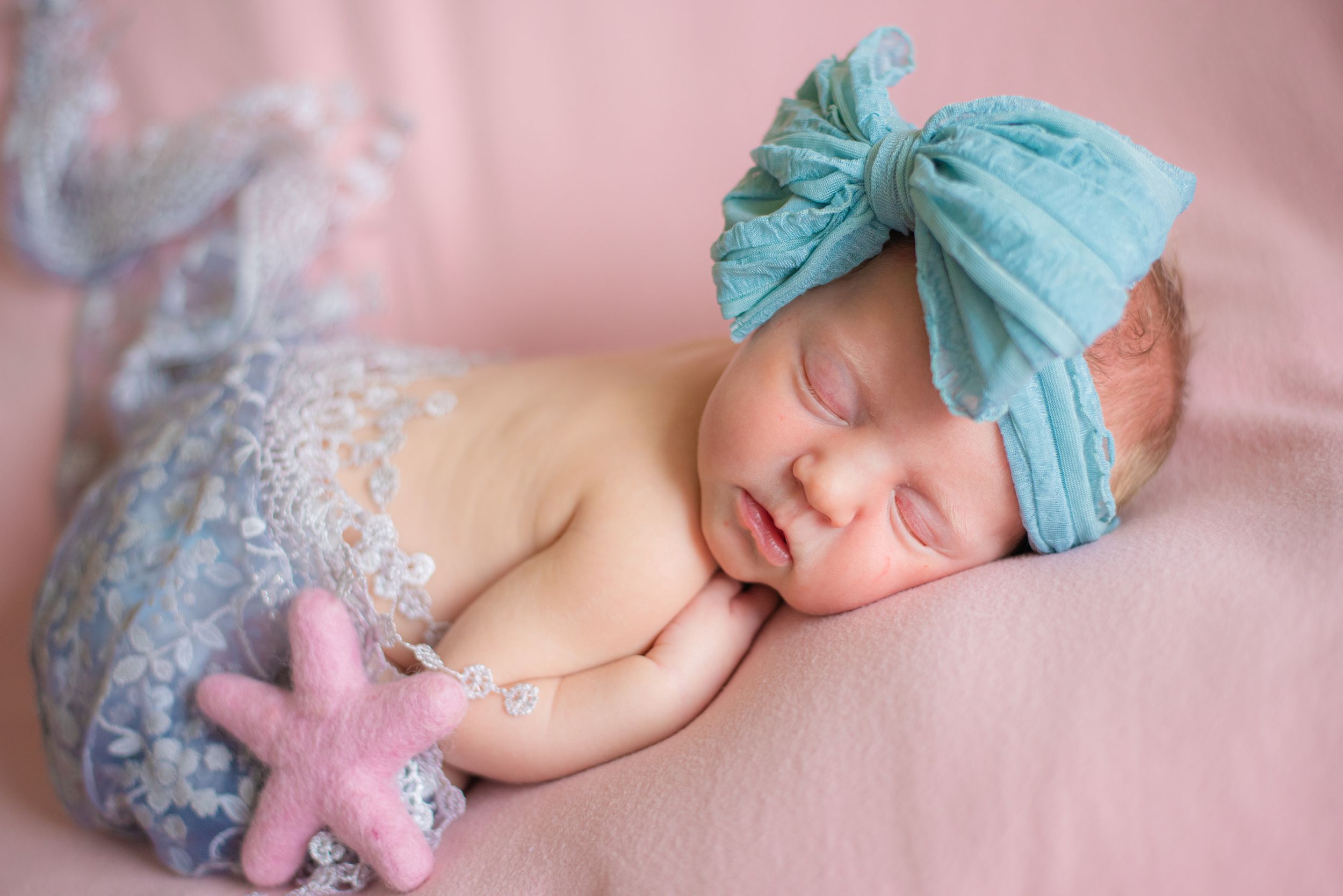 Sleeping newborn with turquoise bow headband and pink star plush on pink background.