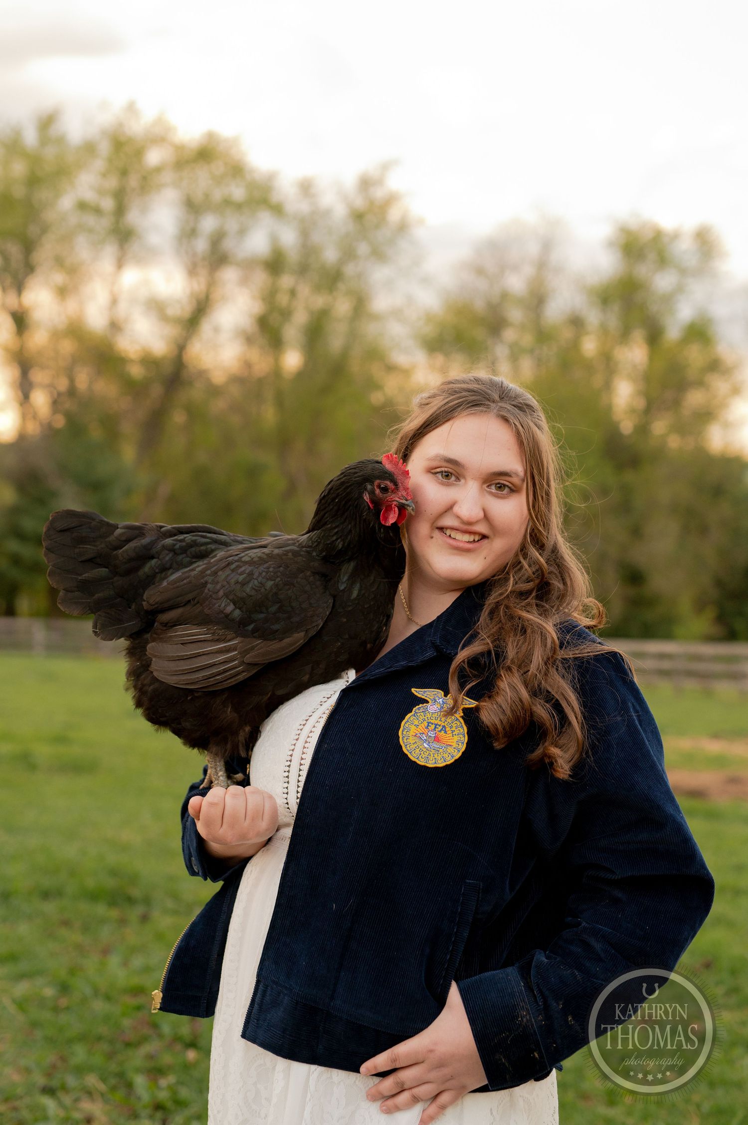 A person in navy jacket poses with a black hen against sunset farm scenery.