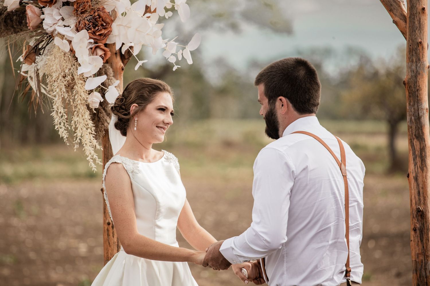 A romantic wedding ceremony under a boho dried flower arch in a rustic outdoor setting.