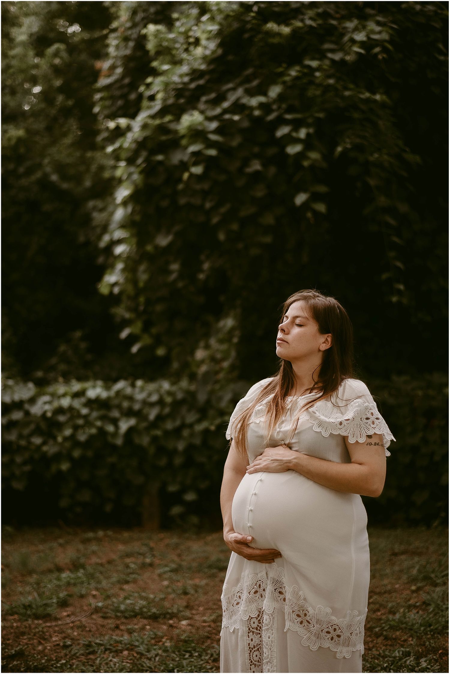 Artistic maternity photo of pregnant woman in white dress with eyes closed against ivy wall.