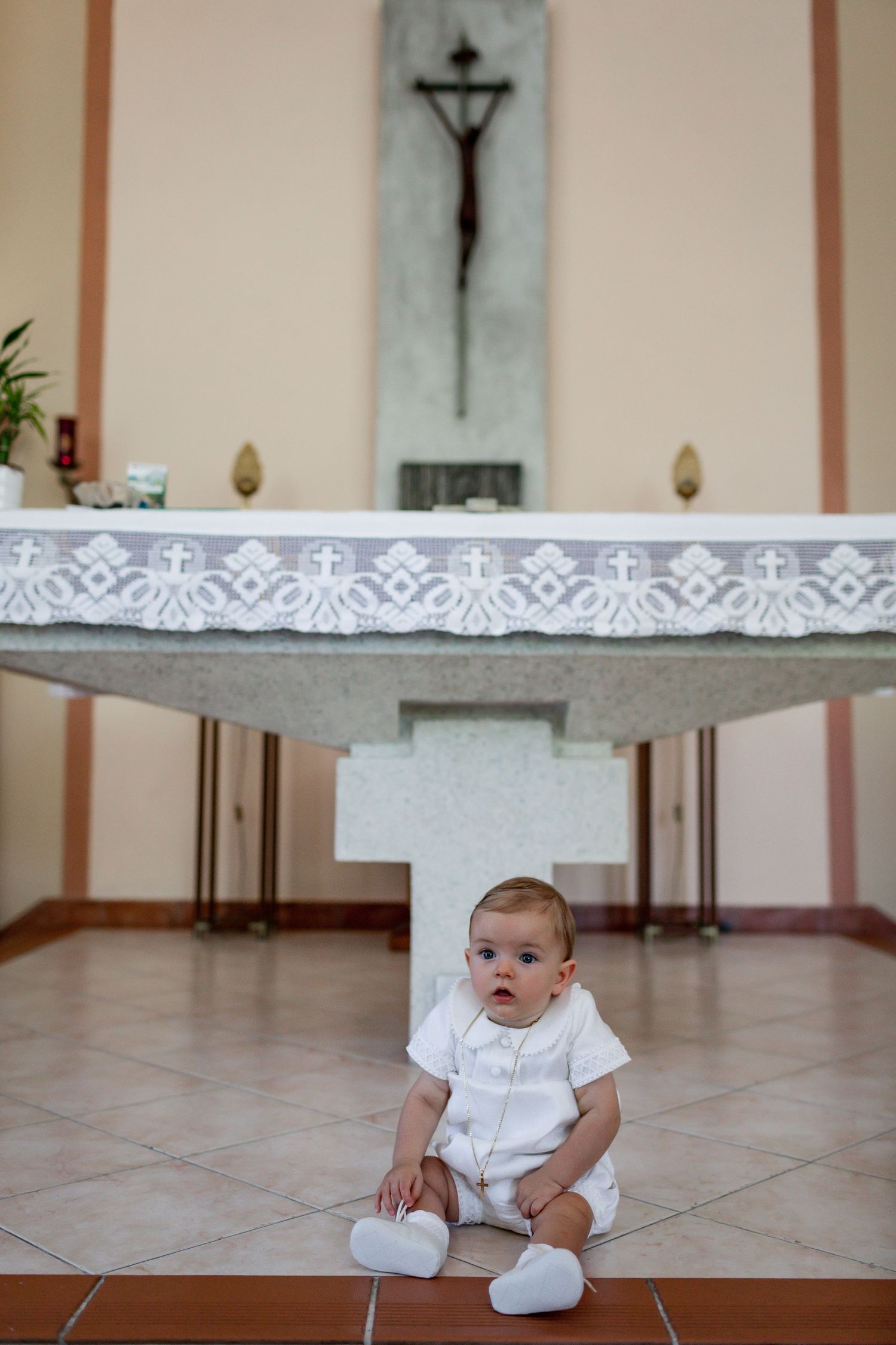 A baby in white christening attire sits beneath an altar with lace details.