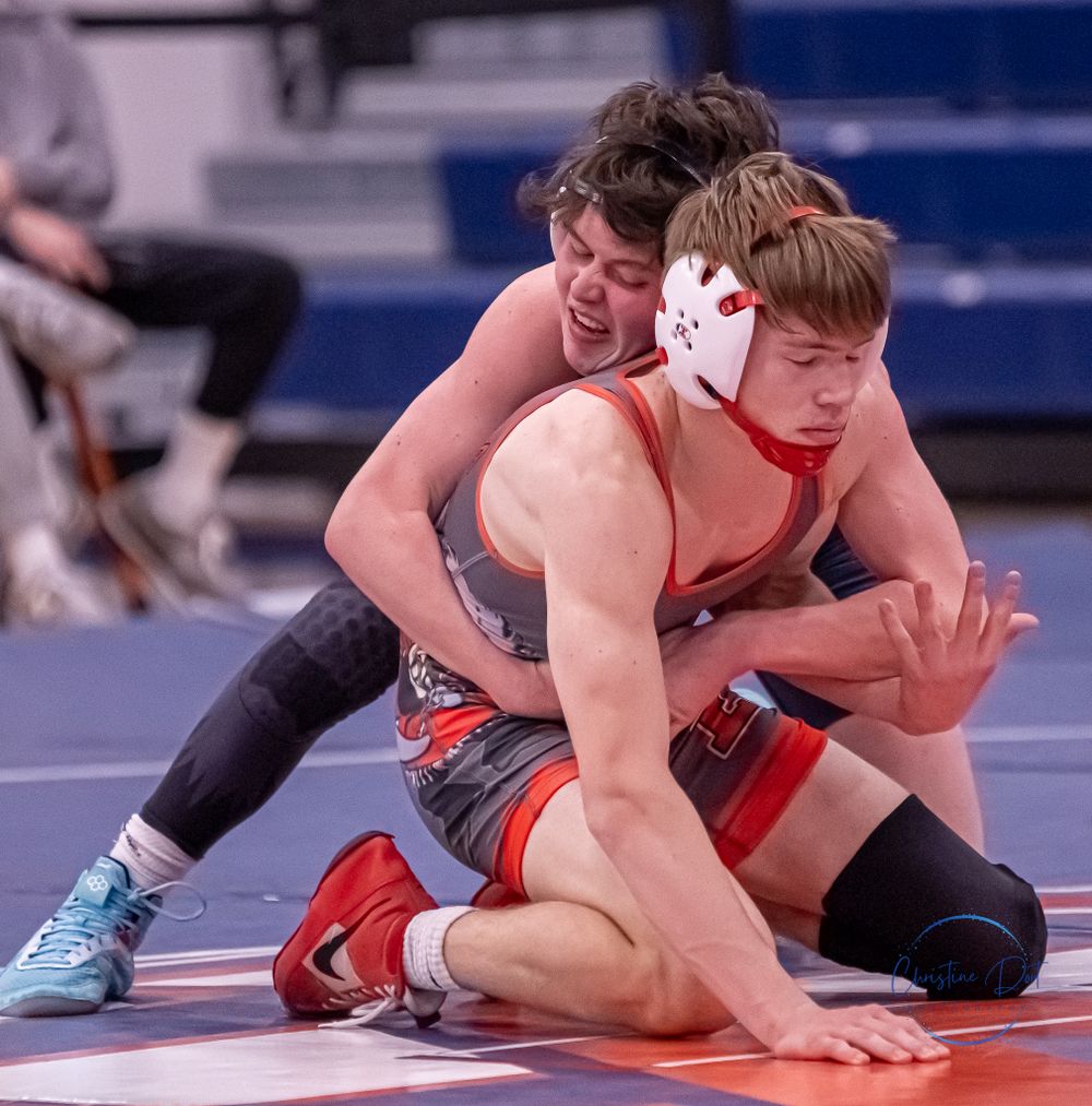 Two wrestlers compete on a blue mat during an intense wrestling match with one wearing protective headgear.