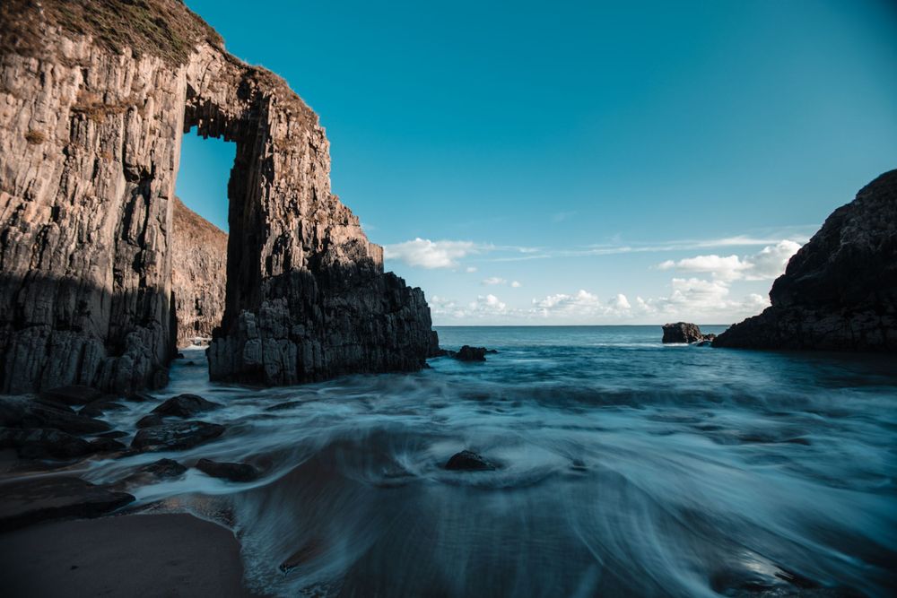 Natural rock arch formation over turbulent ocean waters against bright blue sky.