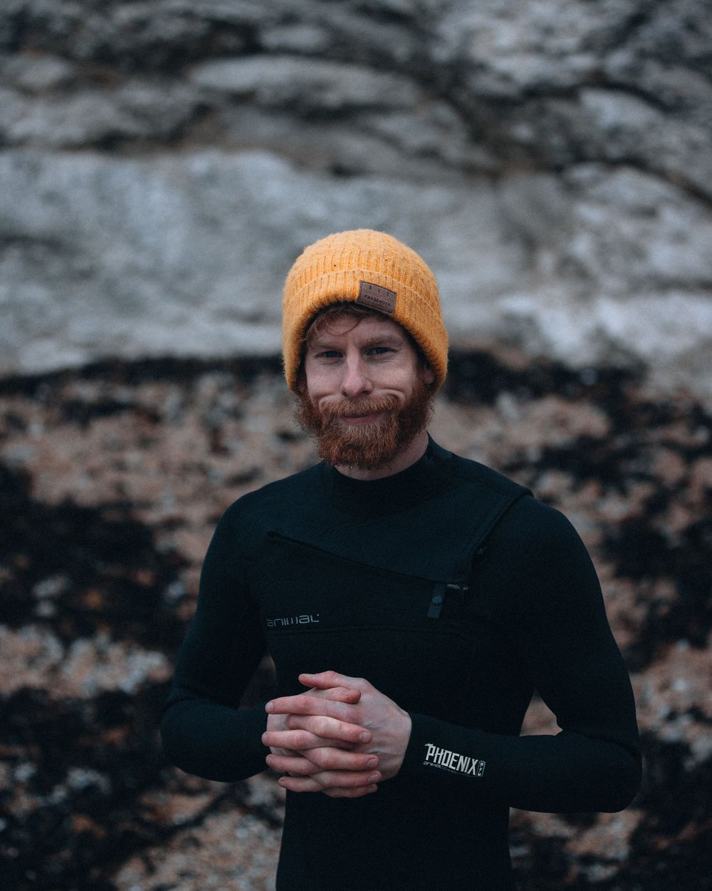Person wearing a yellow beanie and black wetsuit against rocky backdrop.