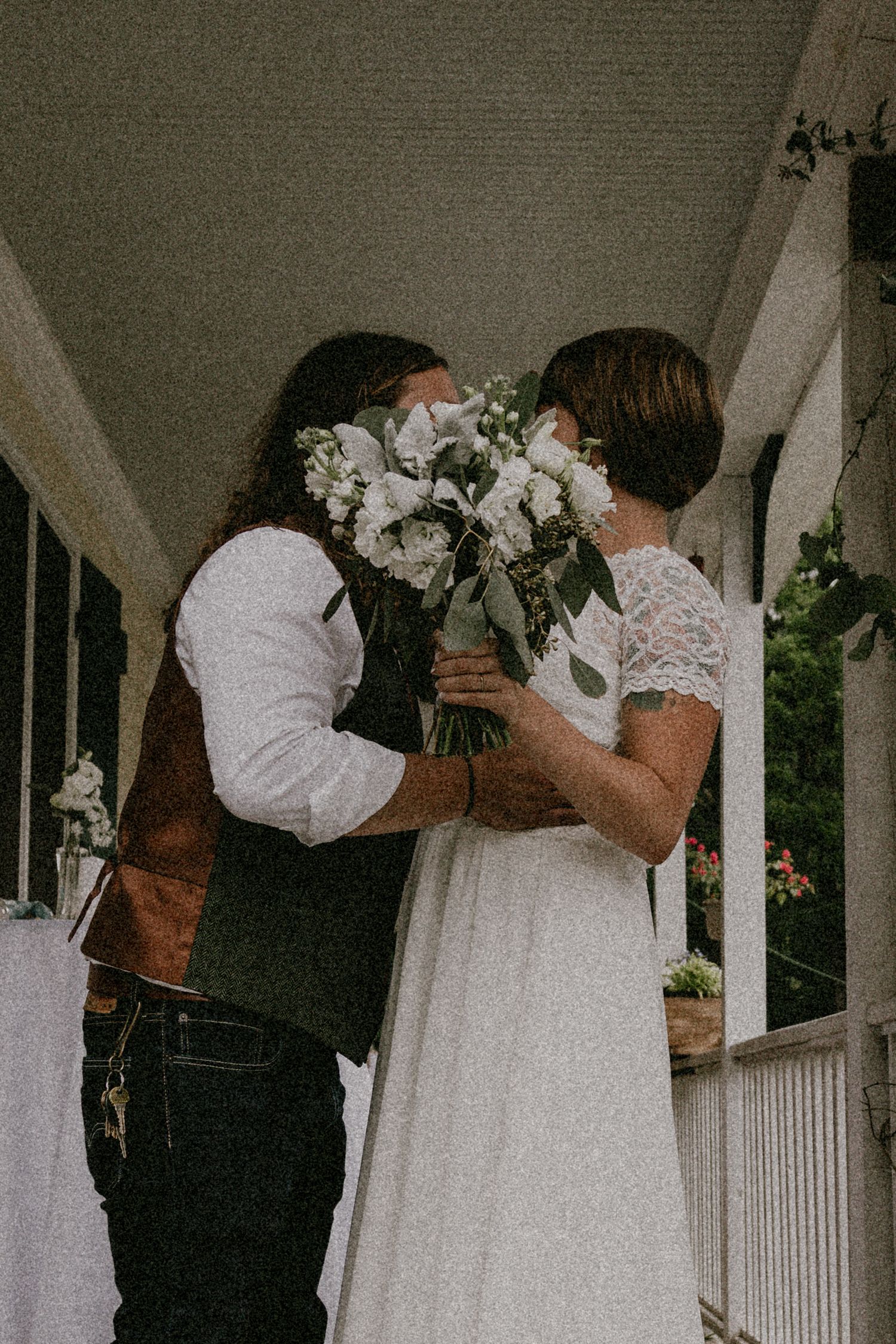 A romantic wedding couple sharing a tender moment behind a bouquet of white flowers on a porch.