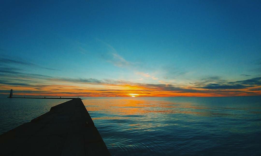 Vibrant orange sunset over calm water with a dark rocky jetty in the foreground.