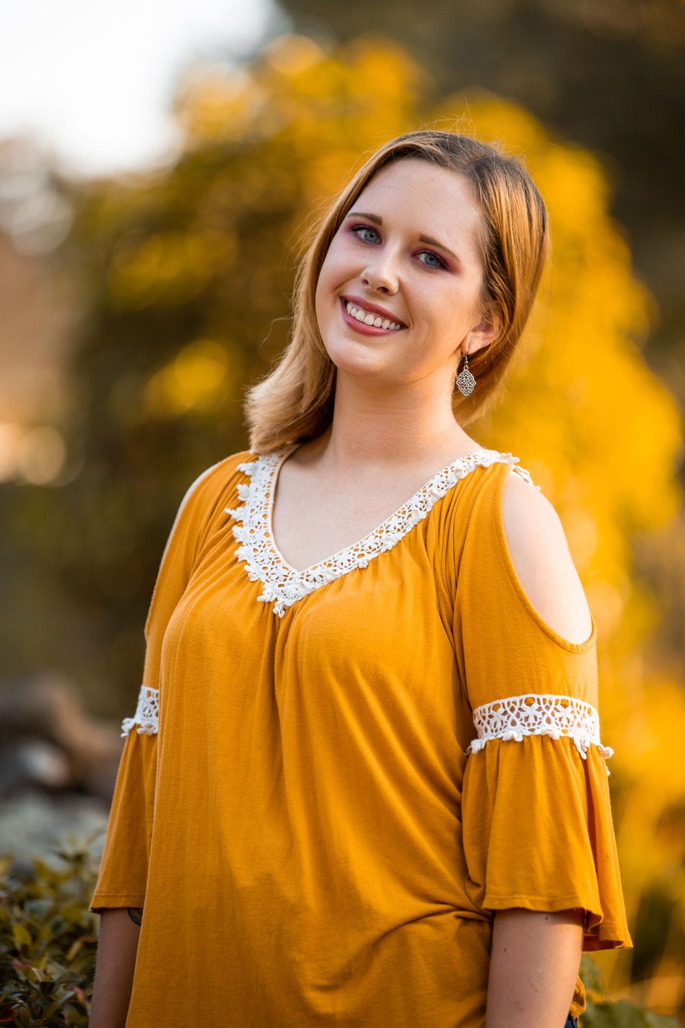 Portrait in mustard yellow cold-shoulder top against autumn foliage background.