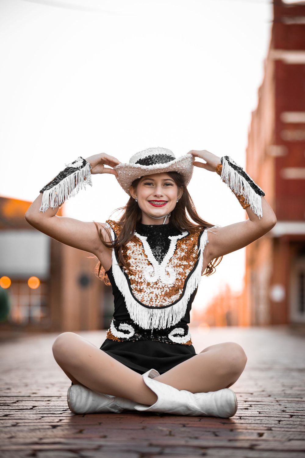 Performer in western costume sitting cross-legged and adjusting hat outdoors.