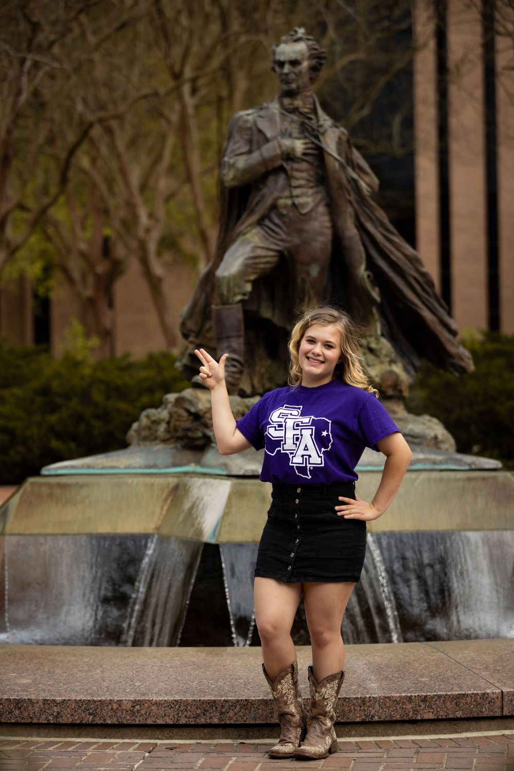 A casual portrait in front of a bronze statue fountain wearing western boots and university apparel.