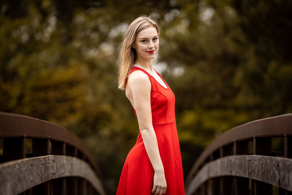 A person in a vibrant red dress poses gracefully on a wooden bridge surrounded by autumn colors.