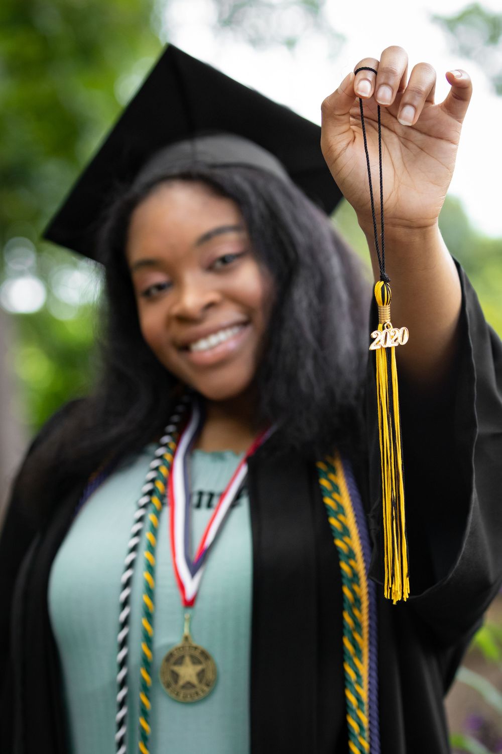 Graduate holding up gold tassel with 2020 charm against blurred green background.