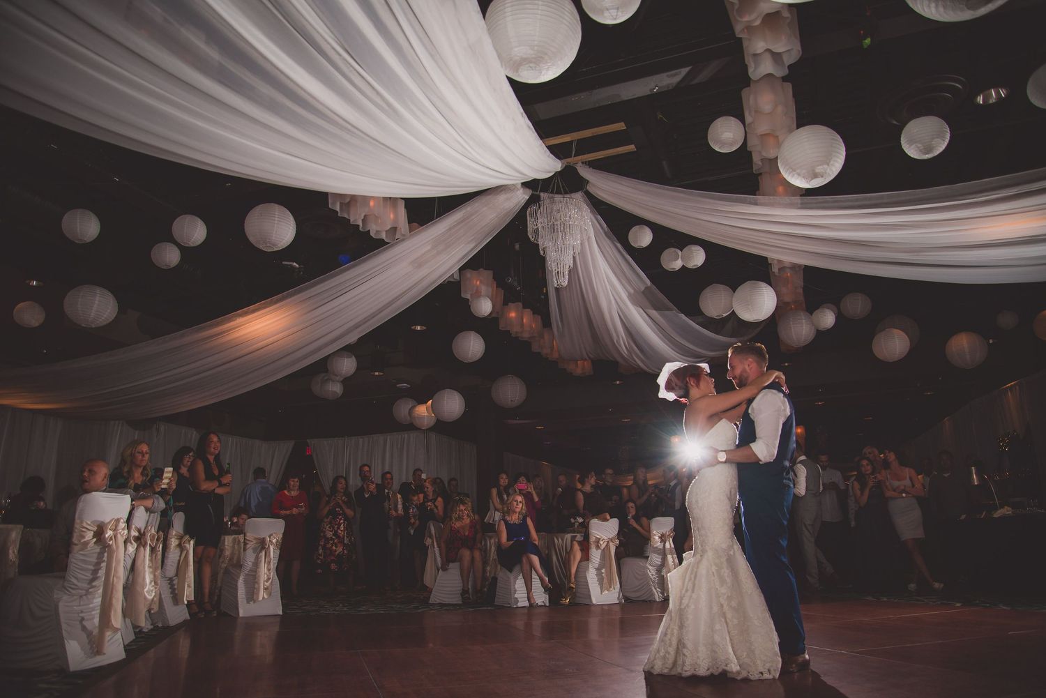 Newlyweds share first dance under elegant white draping and paper lantern ceiling decorations.
