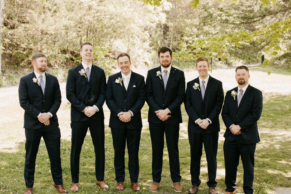 Six groomsmen wearing matching navy suits standing together smiling outdoors at a wedding.