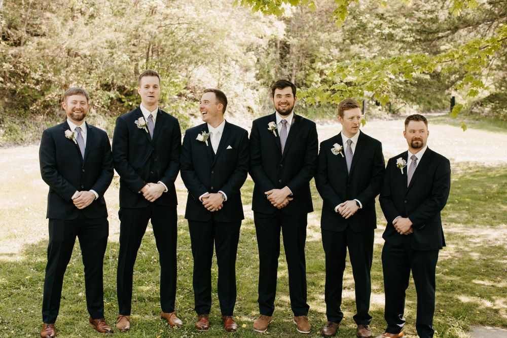 Six groomsmen in navy suits with floral boutonnieres posing outdoors at a wedding ceremony.