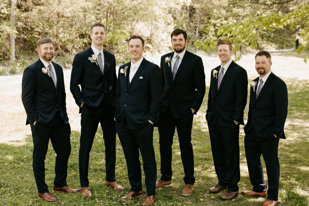 Six groomsmen in dark navy suits posing casually with hands in pockets at outdoor wedding.