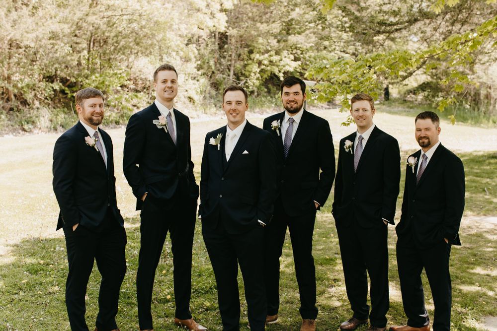 Groom and groomsmen in navy suits with gray ties laughing together on green lawn at wedding.