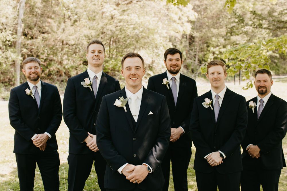 Groom in navy suit with boutonniere stands before five groomsmen in outdoor wedding portrait.
