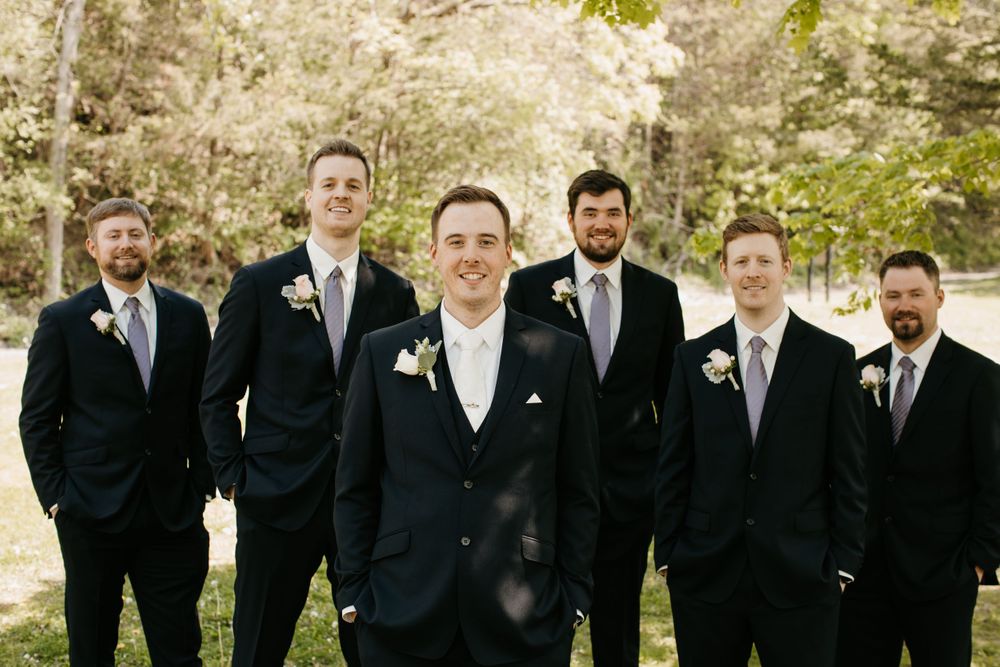 Groom stands in front of groomsmen in navy suits posing for outdoor wedding party photo.