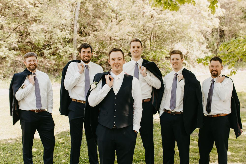 Groomsmen in white shirts holding navy suit jackets over shoulders with groom at wedding.