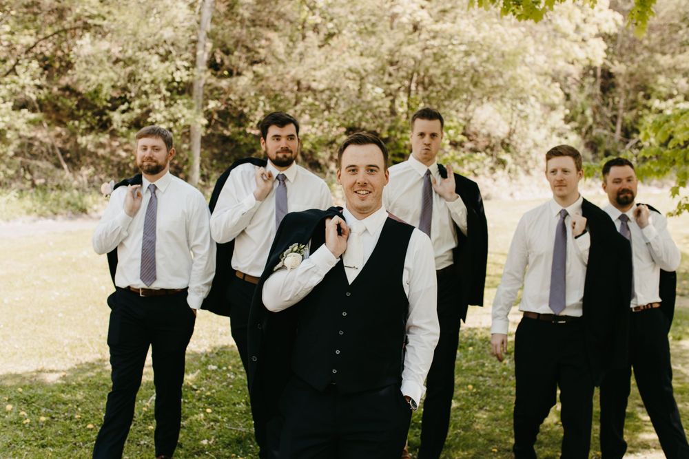 Groom in navy vest leads groomsmen holding jackets in fun outdoor wedding party portrait.